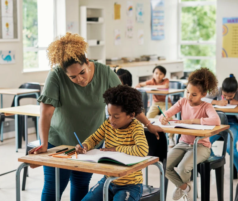 Teacher helps an elementary age boy sitting at a desk in a classroom of students.