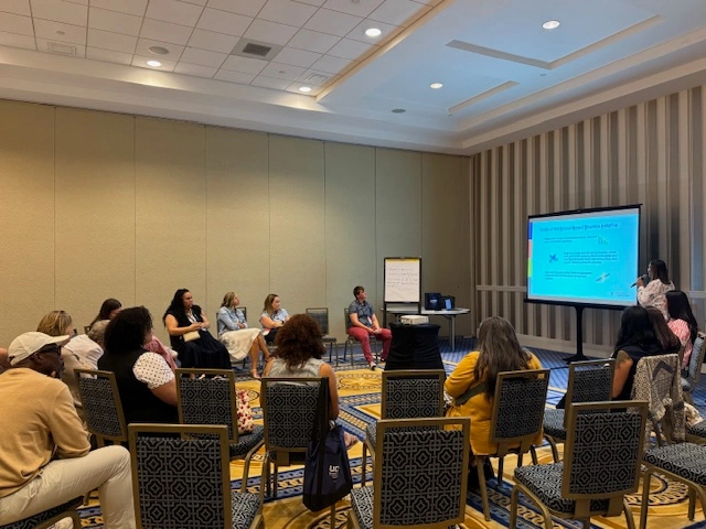 A group of about 20 adults sit in chairs in a circle in a large conference room