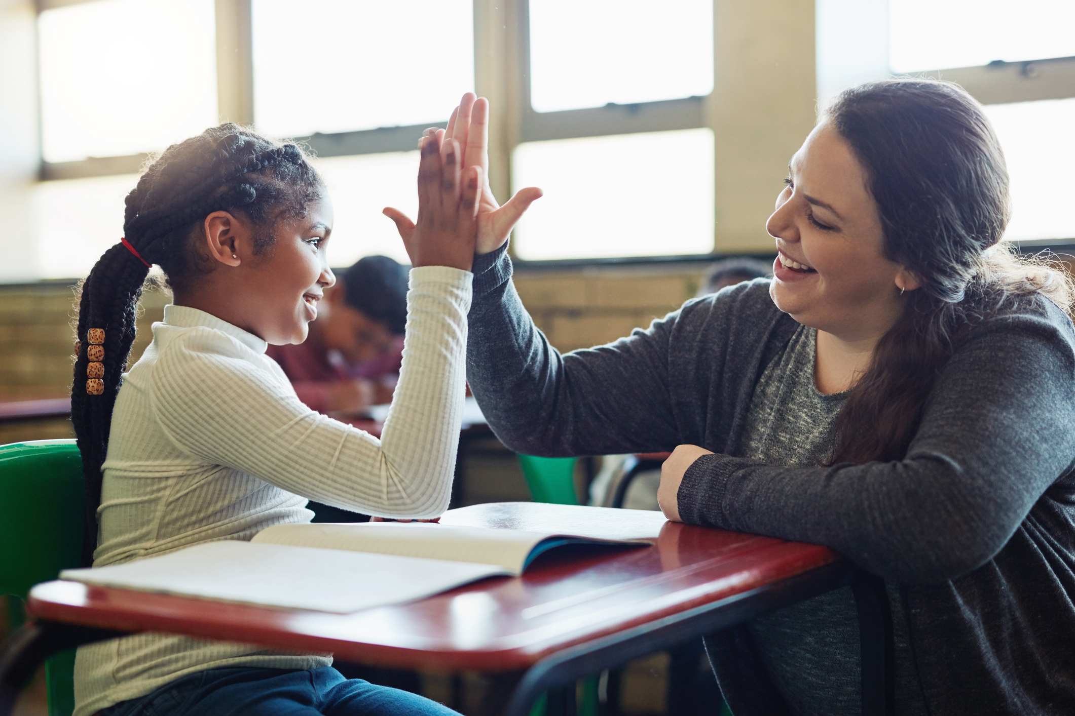 Middle school aged girl and teacher sit together at a desk in a classroom, smiling and high fiving.